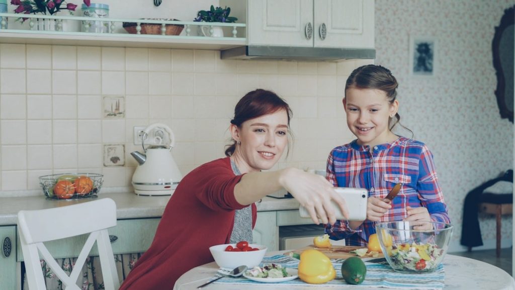 Smiling mother making selfie portrait together with young cute daughter cooking breakfast at home in kitchen. Family, cook, and people concept
