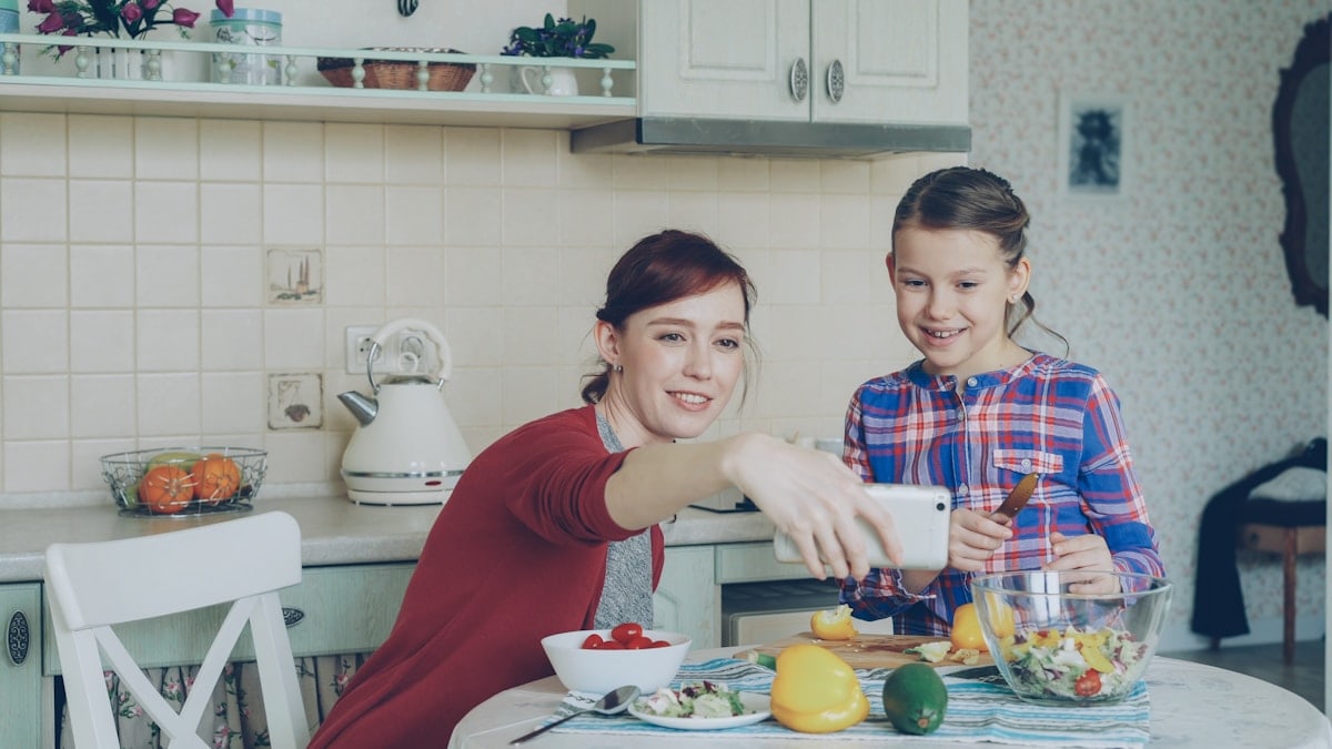 Smiling mother making selfie portrait together with young cute daughter cooking breakfast at home in kitchen. Family, cook, and people concept